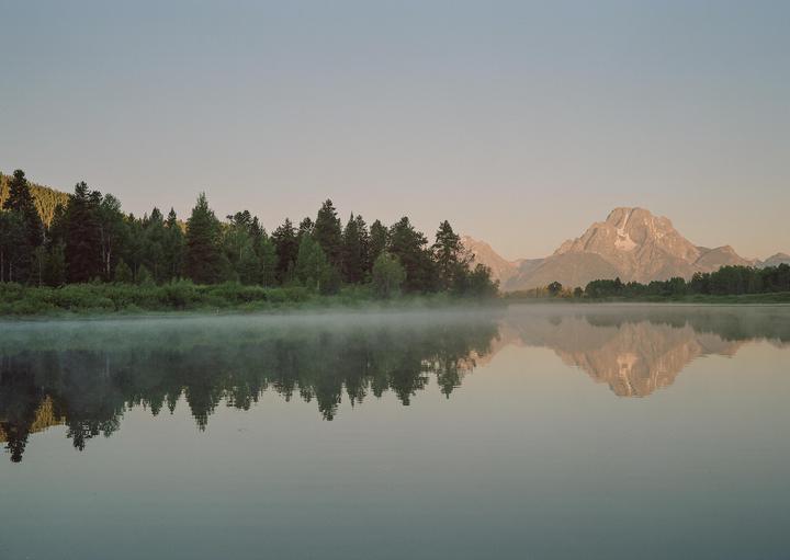 A misty lake reflects a forest and mountain under a clear sky.