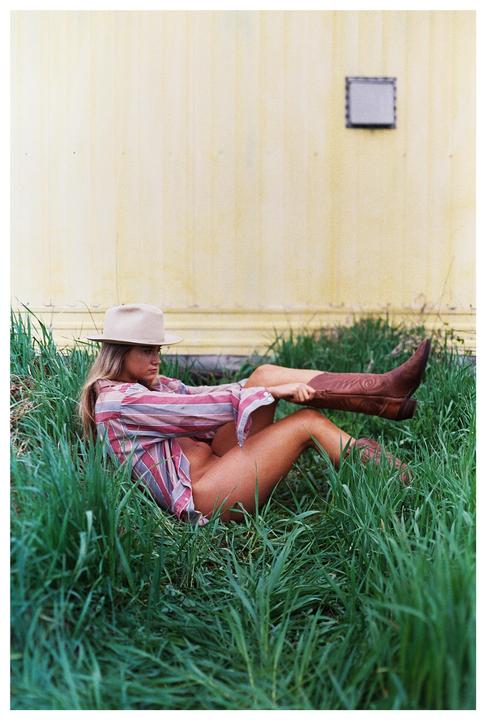 A woman wearing a cowboy hat, a striped shirt, and cowboy boots is lying in tall grass next to a yellow wall.