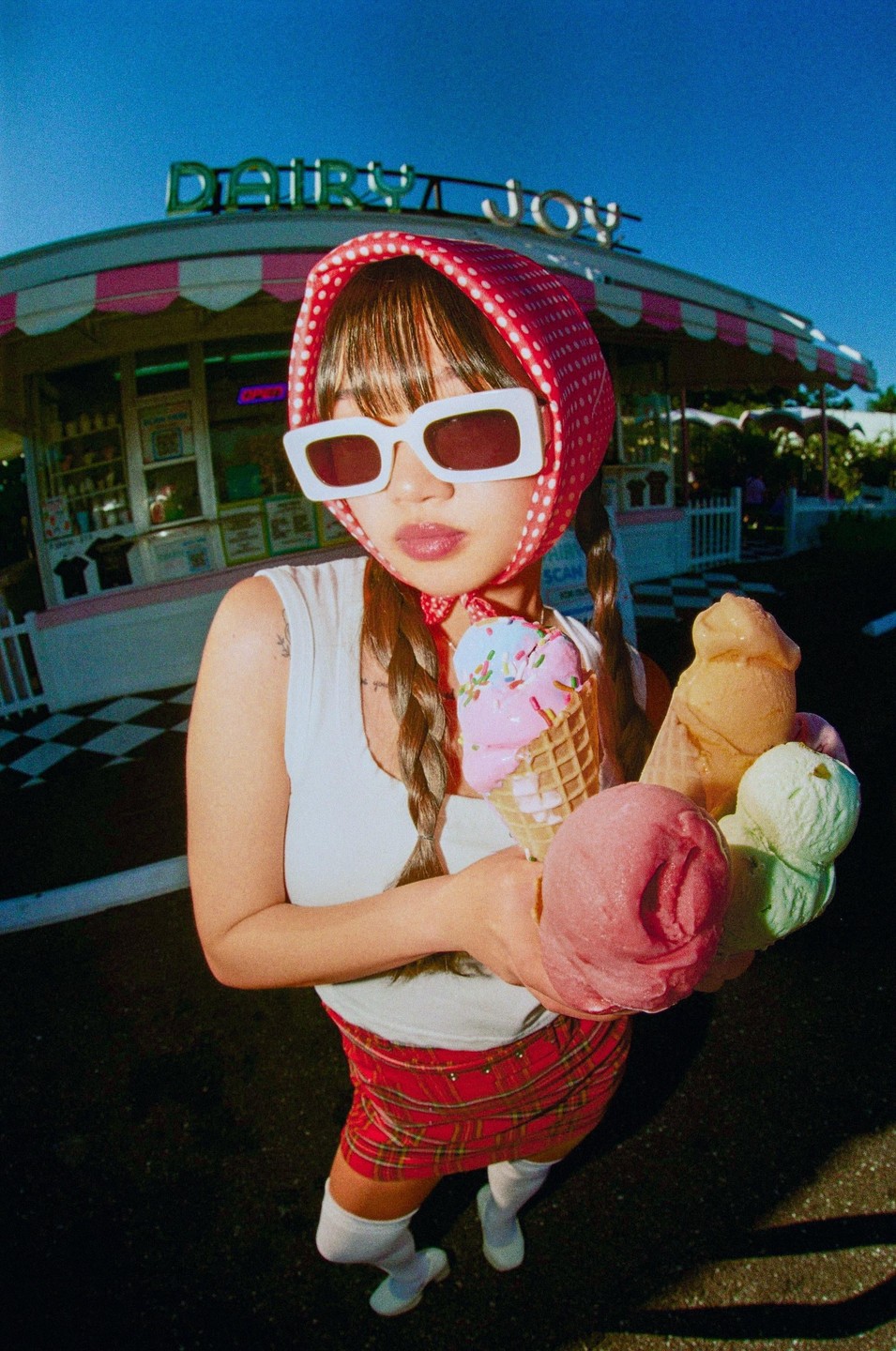 A woman in a red headscarf and sunglasses holds multiple ice cream cones in front of an ice cream parlor.