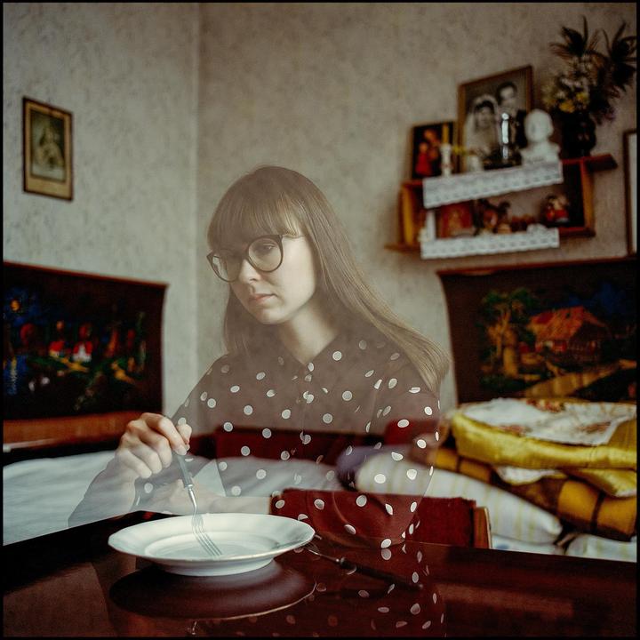 A woman wearing glasses and a polka dot shirt sits at a table with a plate and fork, with reflections of the room visible.