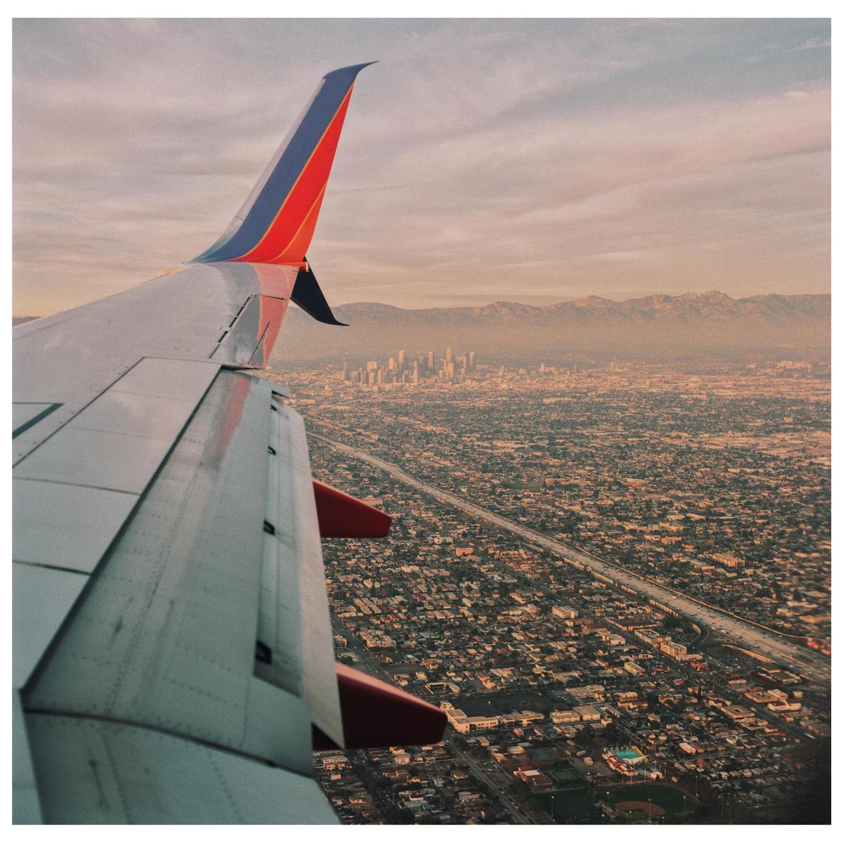 An airplane wing is visible over a sprawling city with mountains in the distance.