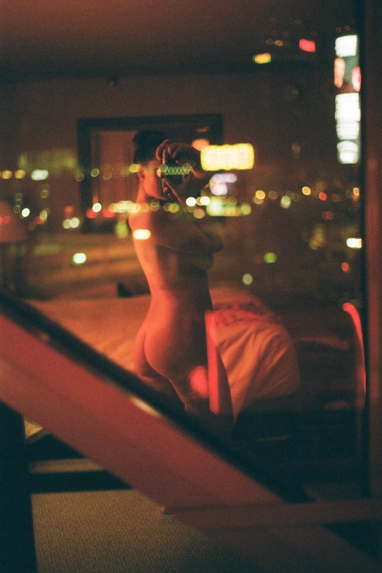 A woman takes a self-portrait in a window reflecting city lights at night.