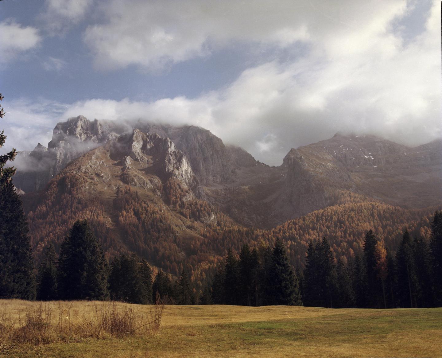 Clouds partially obscure a rugged mountain range with autumn foliage and a line of evergreen trees in the foreground.