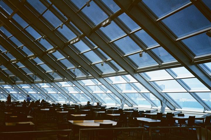A large room with many tables and chairs is illuminated by natural light coming through a glass ceiling and wall.