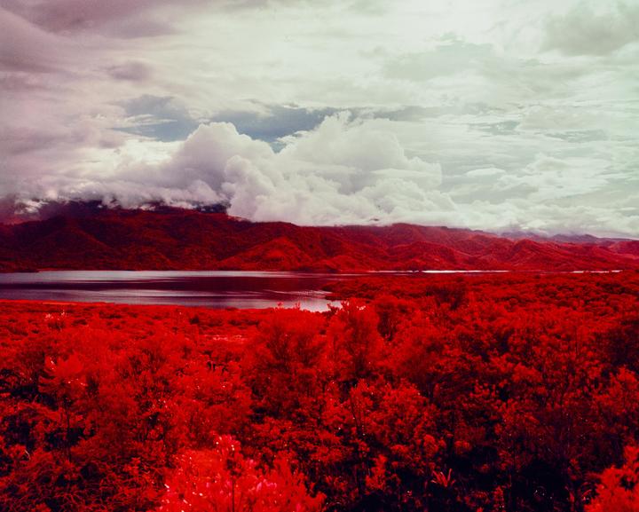A body of water is surrounded by red foliage and mountains under a cloudy sky.