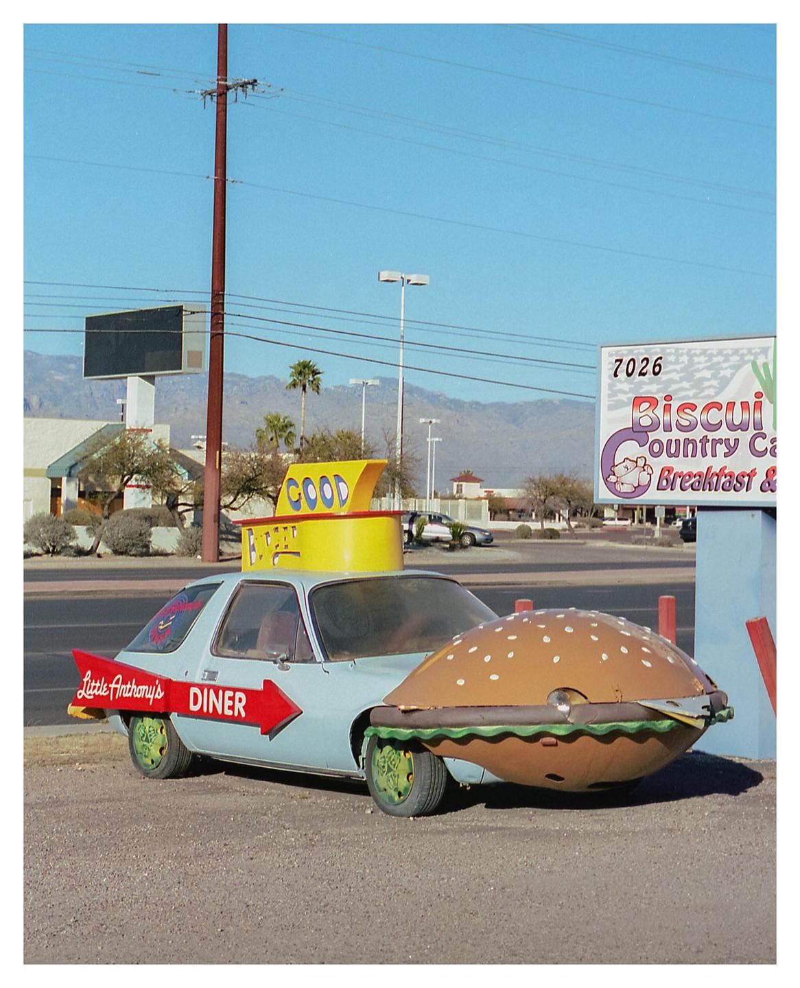 A light blue car with a hamburger-shaped front and a diner sign on the side is parked in front of a sign for Biscuit Country Cafe.