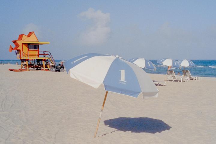 A blue and white beach umbrella casts a shadow on the sand with a lifeguard stand and ocean in the background.