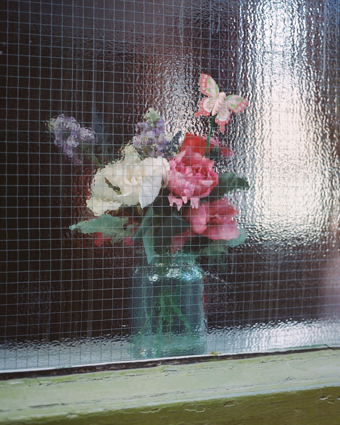 A bouquet of flowers in a vase with a butterfly decoration is seen through a textured window pane with a wire grid.