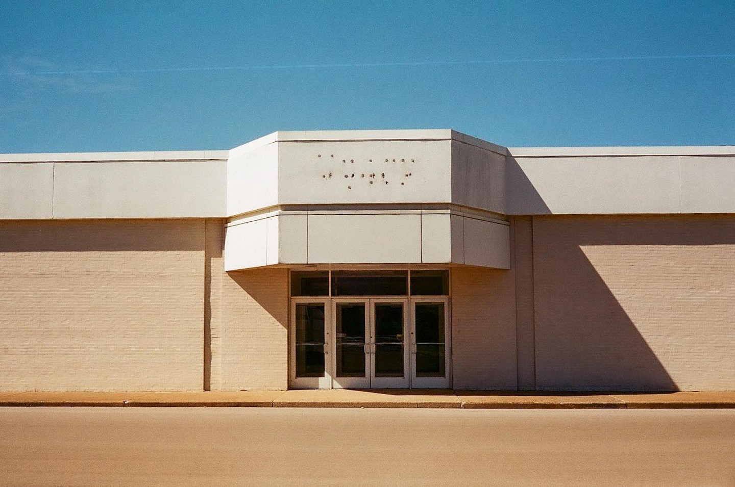 A closed storefront with glass doors and a blank sign is bathed in sunlight, casting sharp shadows on the building's facade.