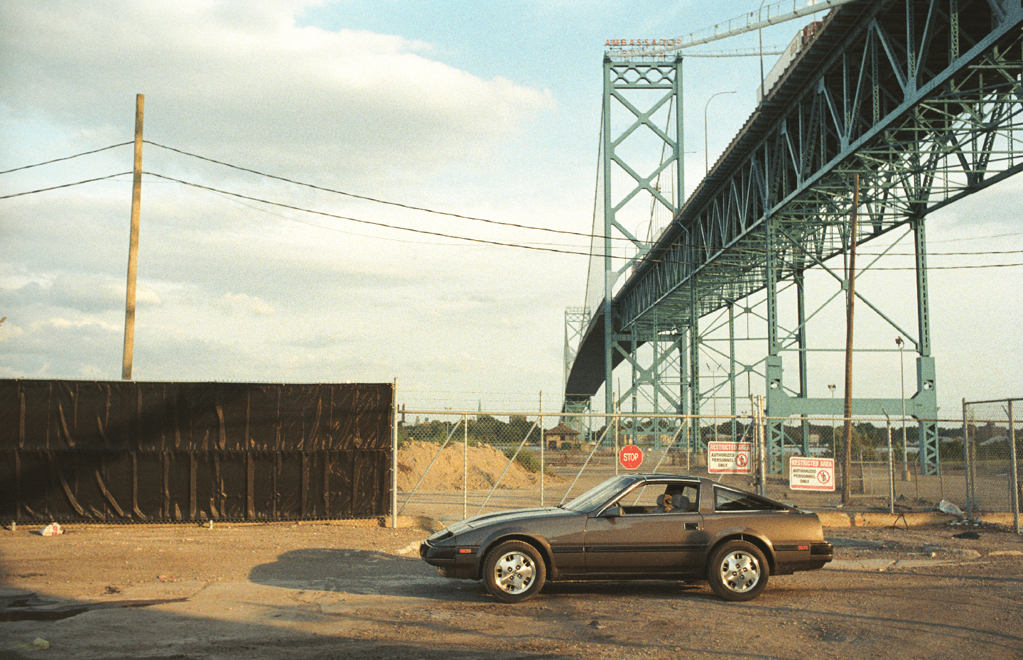 A brown car is parked on a dirt lot next to a fence with a large bridge in the background.