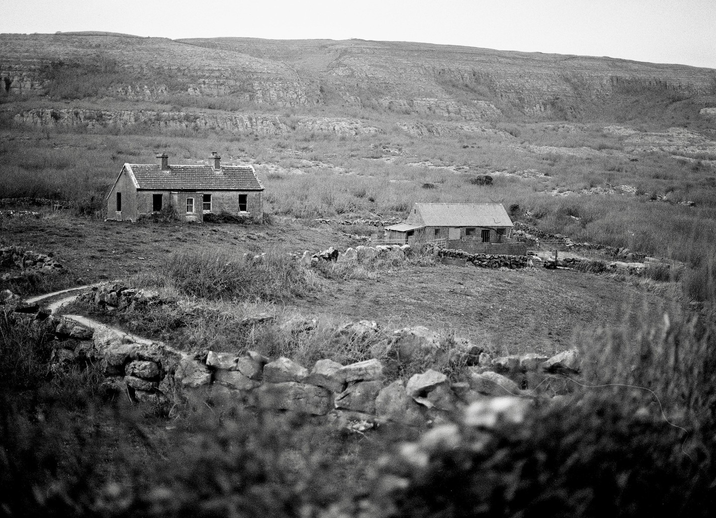 Two abandoned farmhouses sit in a rural landscape with a stone wall and a winding path in the foreground.