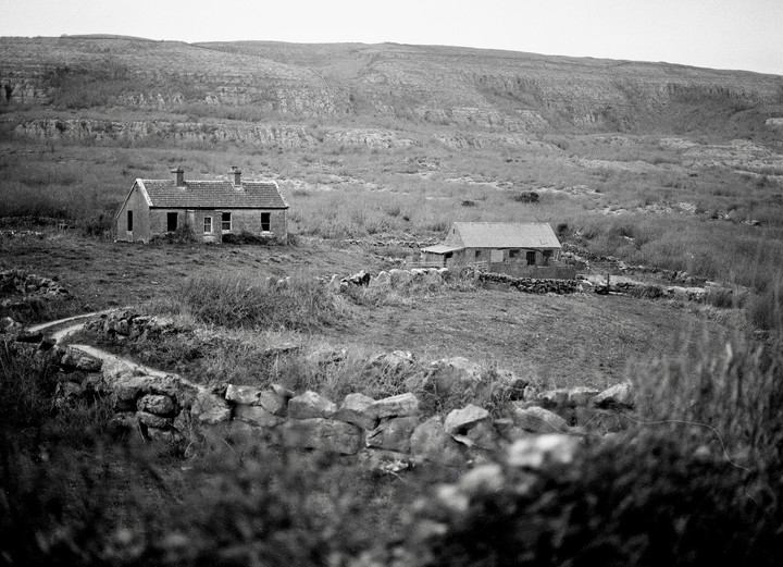 Two abandoned farmhouses sit in a rural landscape with a stone wall and a winding path in the foreground.