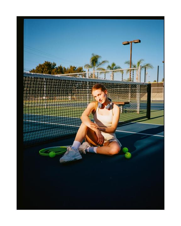 A woman sits on a tennis court next to a tennis racket and tennis balls.