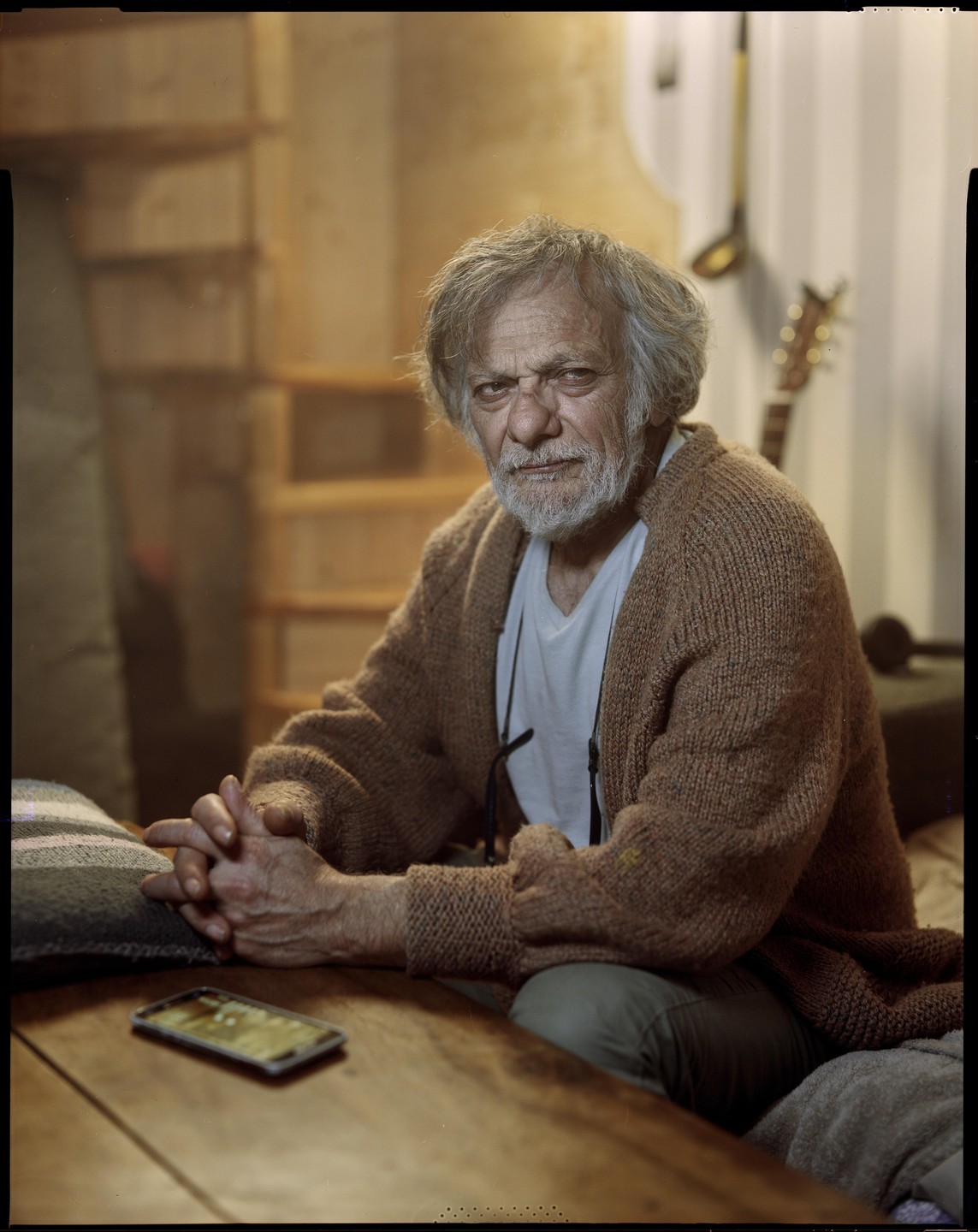 A man with a beard and a brown cardigan sits at a wooden table with his hands clasped.