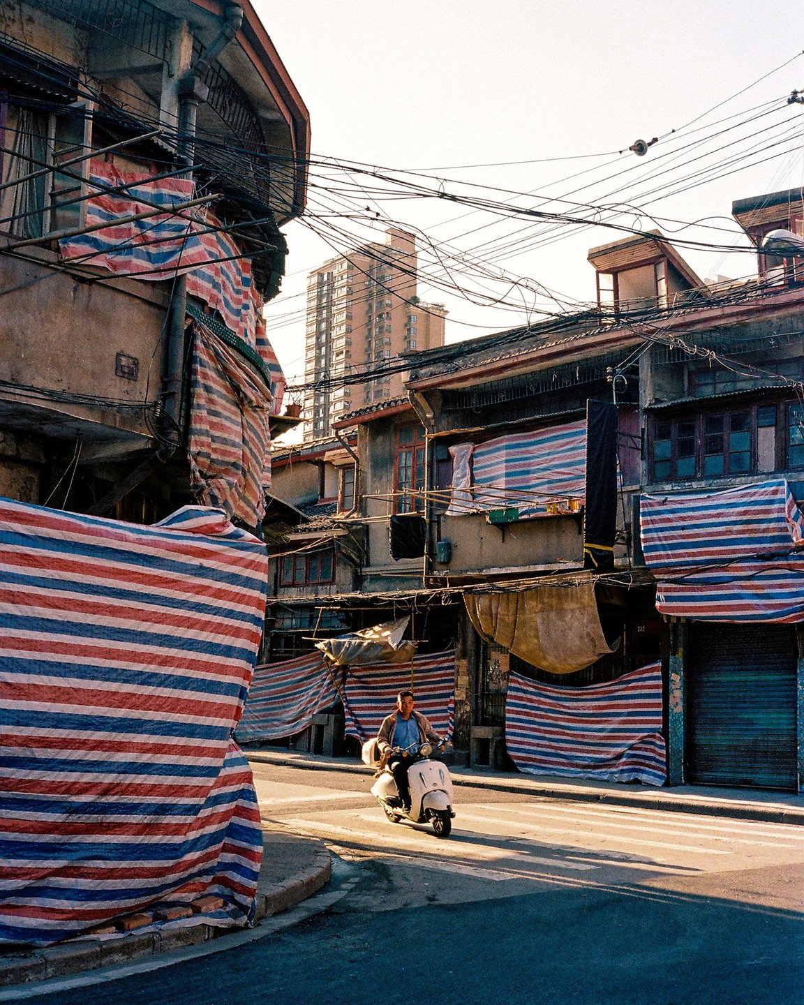A man rides a scooter down a street lined with old buildings covered in striped tarps.