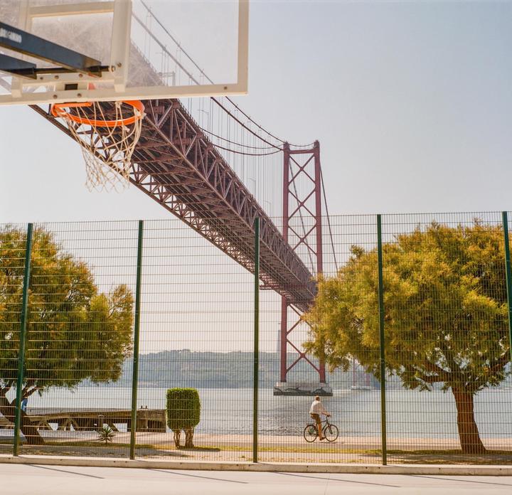 A man rides a bicycle past a fence and trees with a large bridge and body of water in the background.