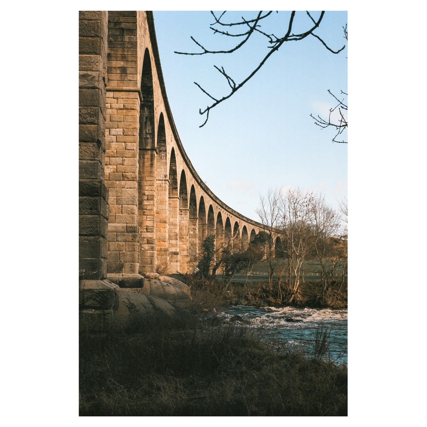 A long stone viaduct with many arches curves over a river and bare trees under a clear sky.