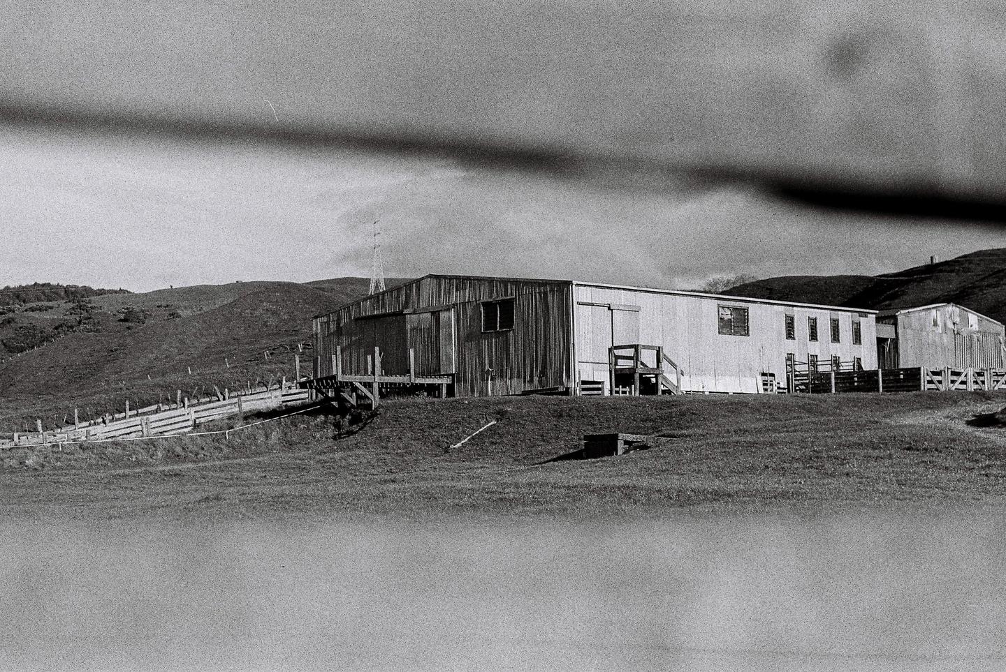 A large wooden woolshed stands on a grassy hill under a cloudy sky.
