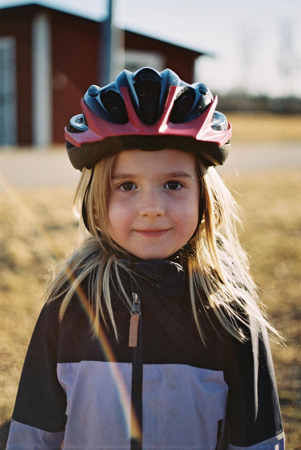 A young girl wearing a red and black bicycle helmet smiles at the camera with a building and field in the background.
