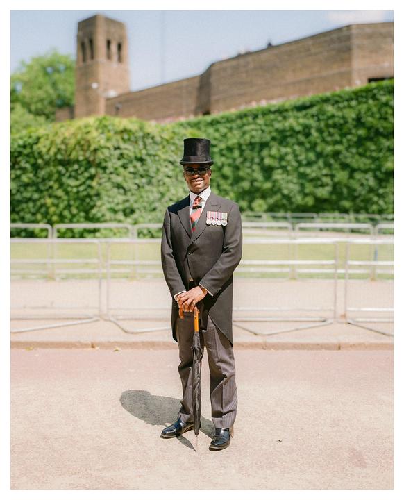 A man in a top hat and formal attire stands holding an umbrella with a building and green foliage in the background.