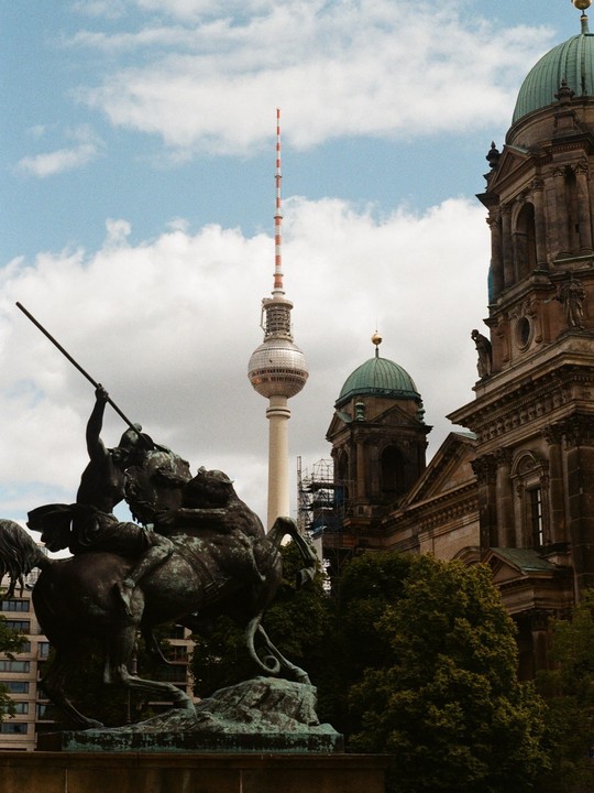 A bronze equestrian statue stands in front of a large building with a green dome and the Berlin TV Tower under a cloudy sky.