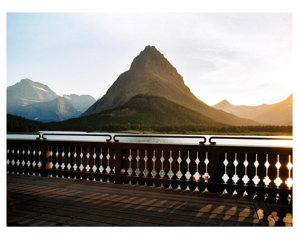 A wooden deck and railing overlook a lake with mountains in the background during golden hour.
