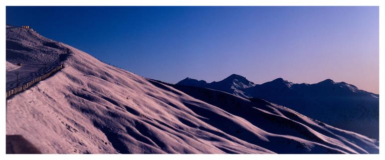 A snow-covered mountain slope with a fence and distant peaks is illuminated by the rising sun under a clear sky.