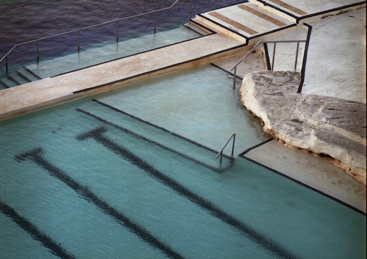 An ocean pool with concrete edges and metal railings is filled with light blue water.