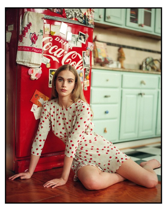 A woman in a polka dot dress sits on a wooden floor next to a red Coca-Cola refrigerator in a kitchen.