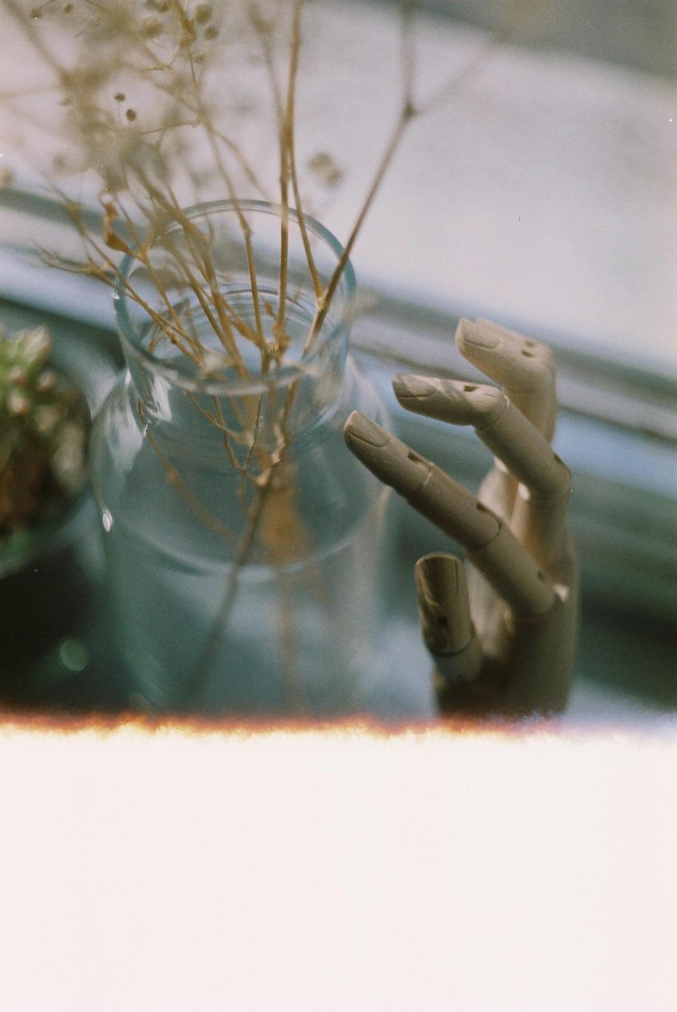A glass vase with dried plants and a wooden mannequin hand are on a window sill.
