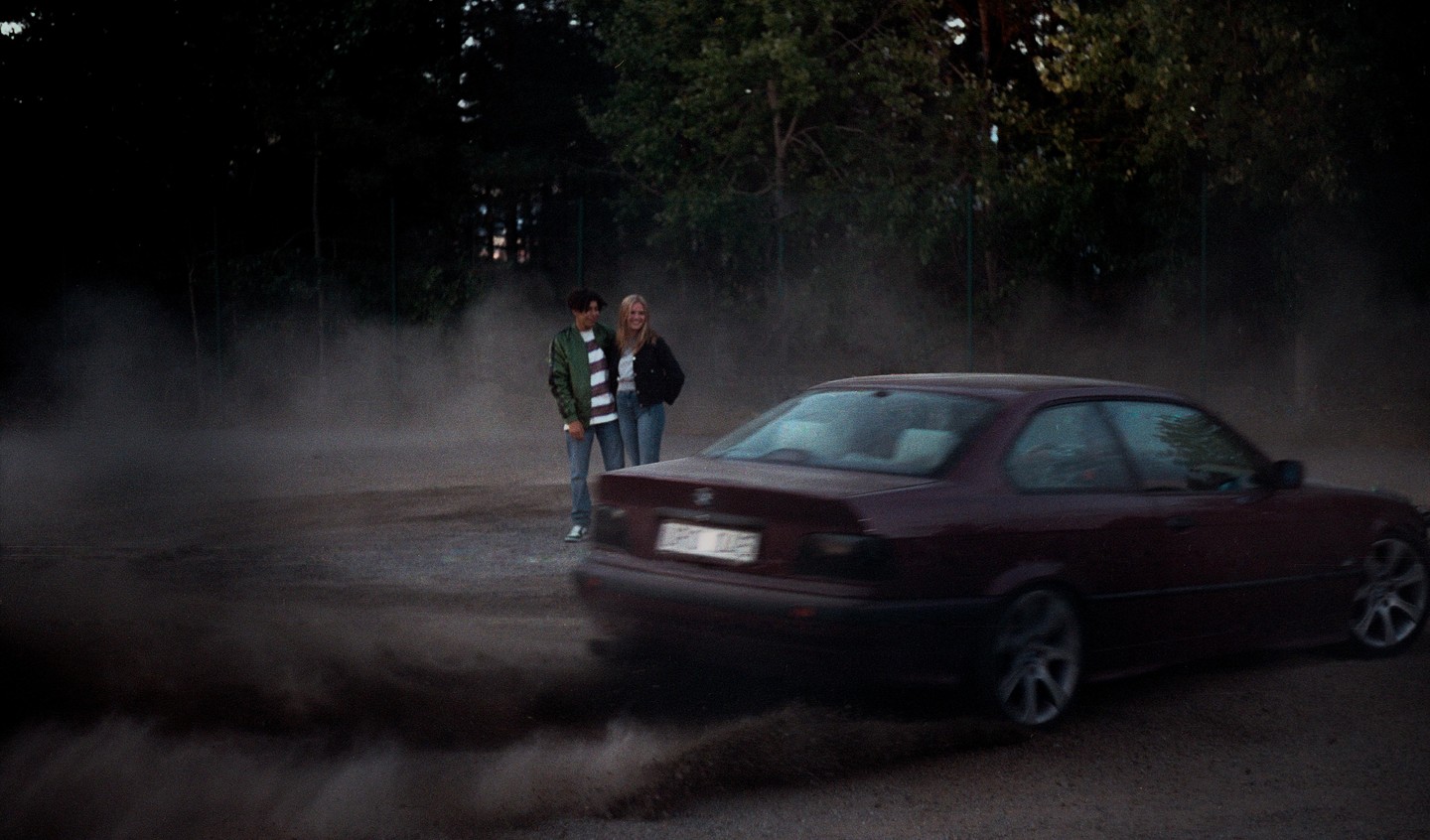 A couple stands in a cloud of dust as a car speeds away from them on a dirt road.