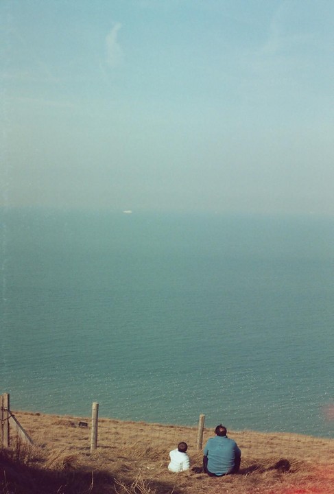 A man and a child sit on a grassy hill overlooking a vast expanse of ocean under a hazy sky.