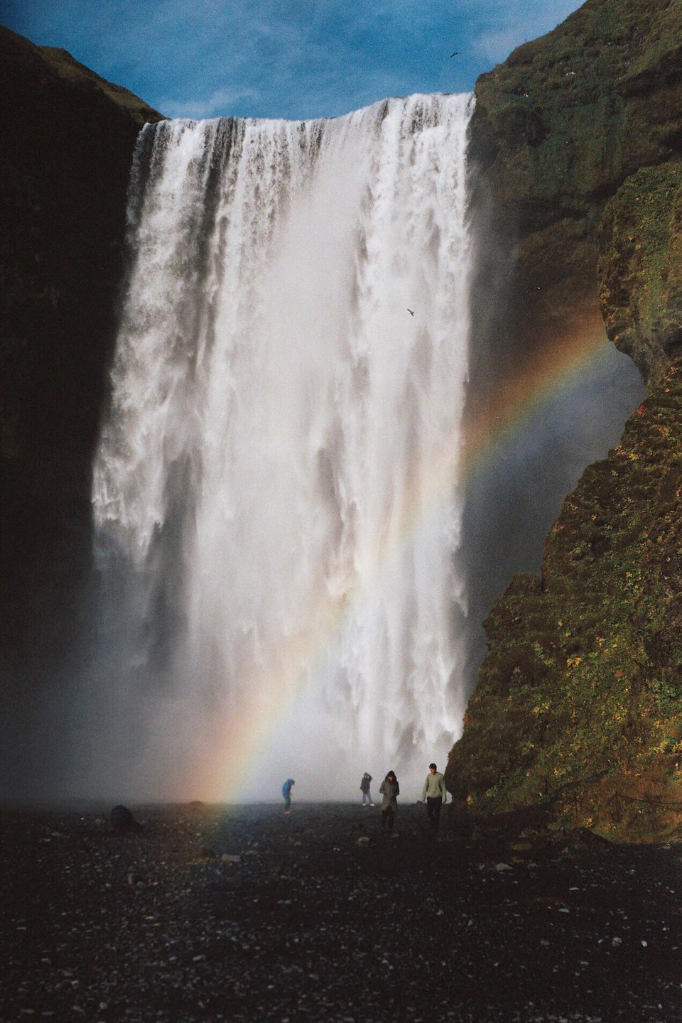 A large waterfall cascades down a cliff face with a rainbow visible at its base, where several people are standing.
