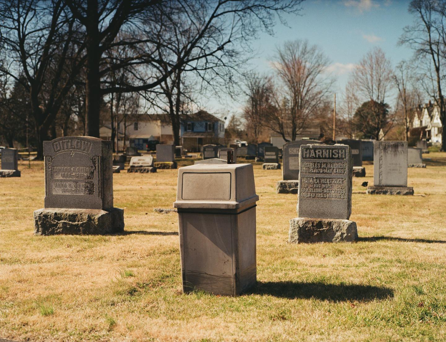 A trash can stands in the middle of a cemetery with gravestones and trees under a clear sky.