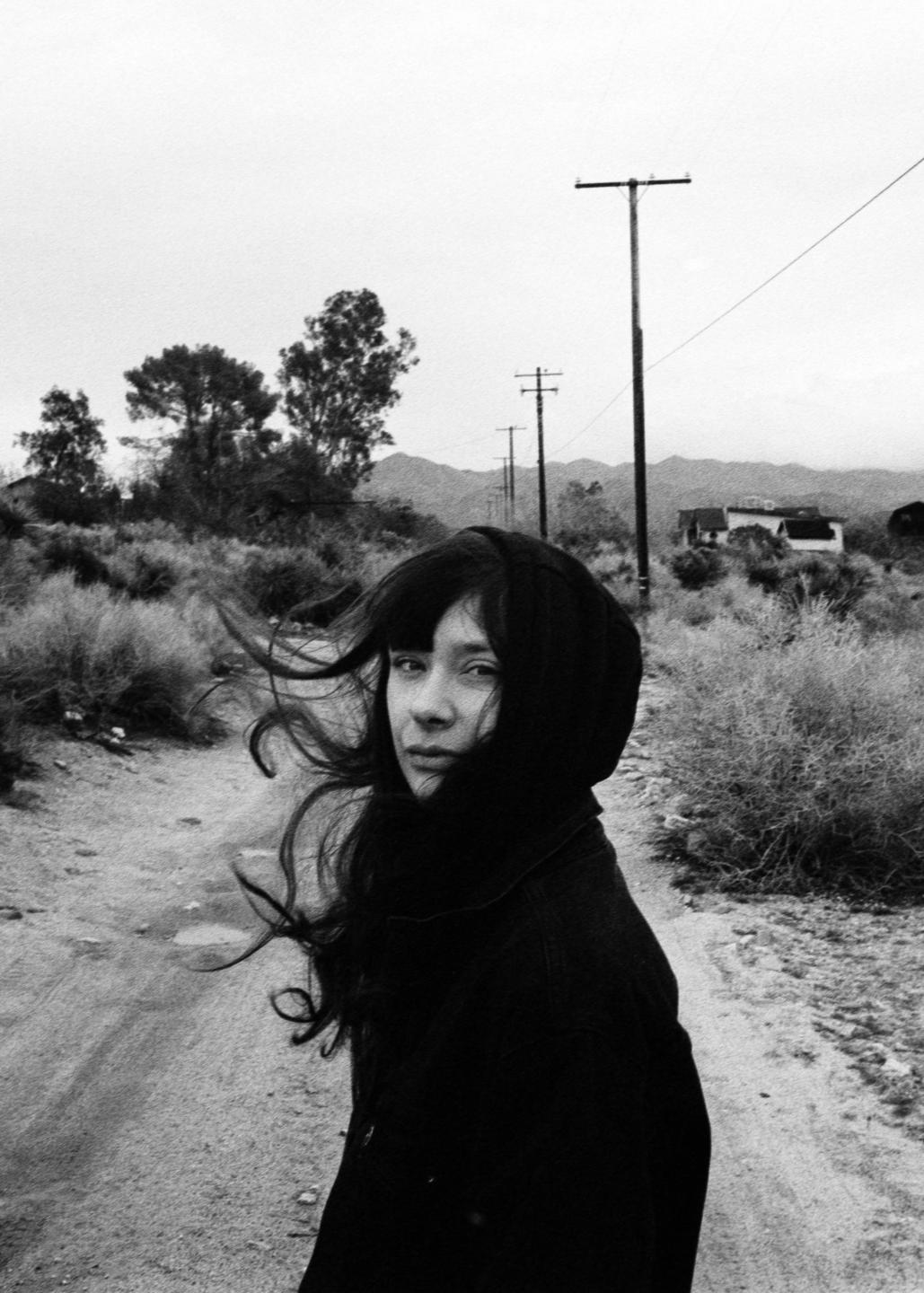 A woman with long, dark hair looks over her shoulder on a dirt road in a desert landscape.