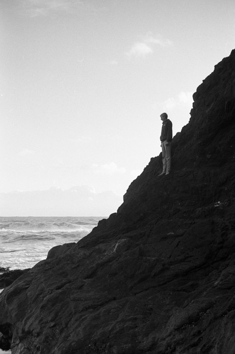 A man stands on a steep, dark rock face overlooking a wavy ocean under a bright sky.