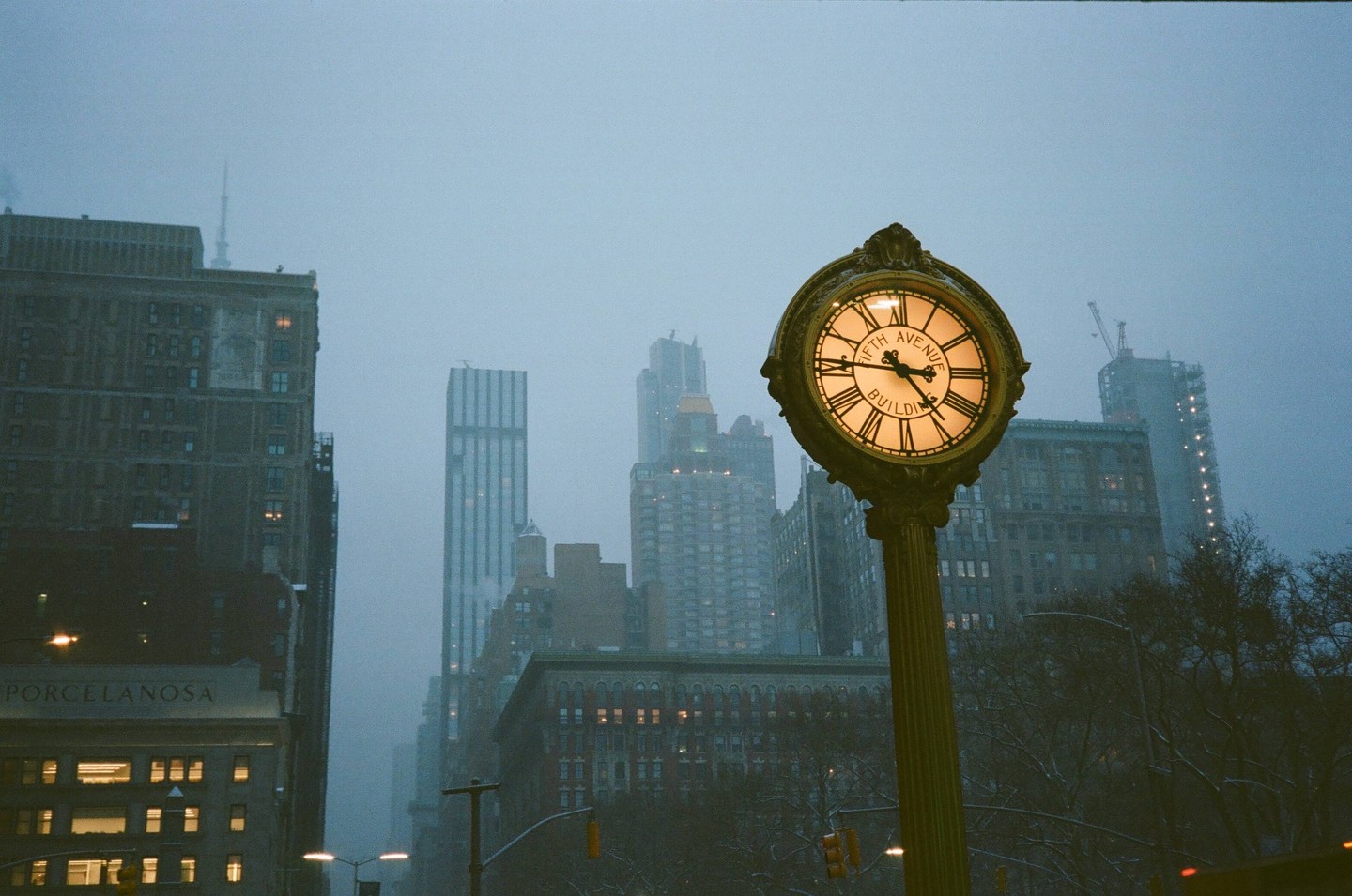 A street clock with "Fifth Avenue Building" written on its face stands in front of tall buildings on a hazy day.