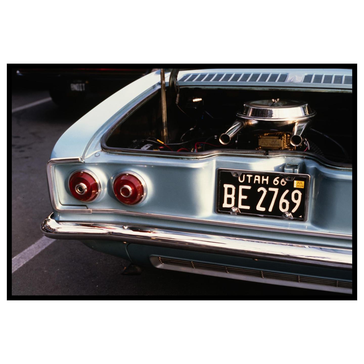 The rear of a light blue vintage car with its trunk open to reveal the engine is parked on asphalt.