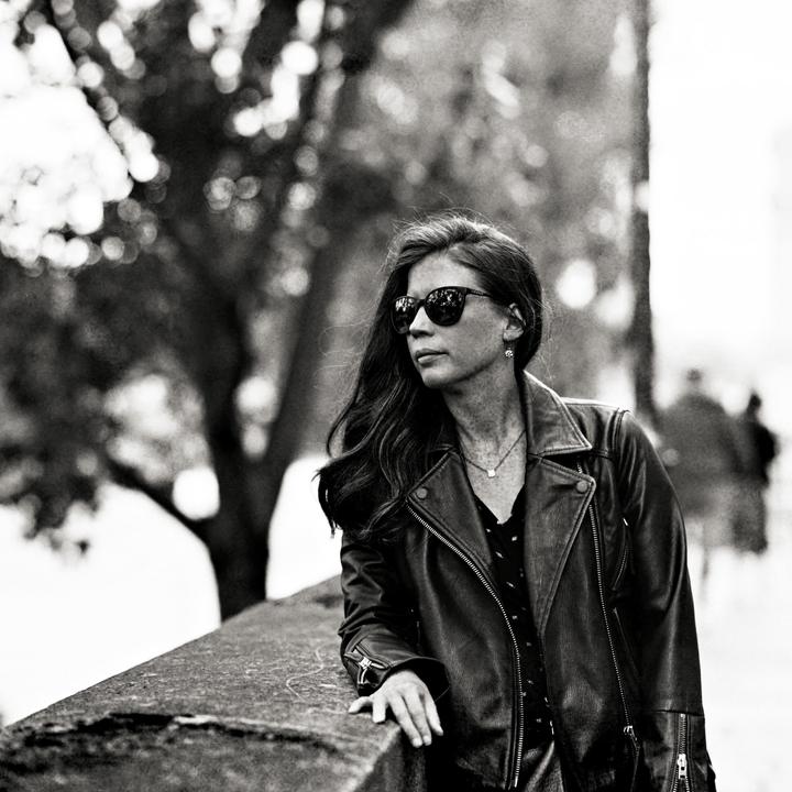 A woman wearing sunglasses and a leather jacket leans on a stone wall with trees in the background.