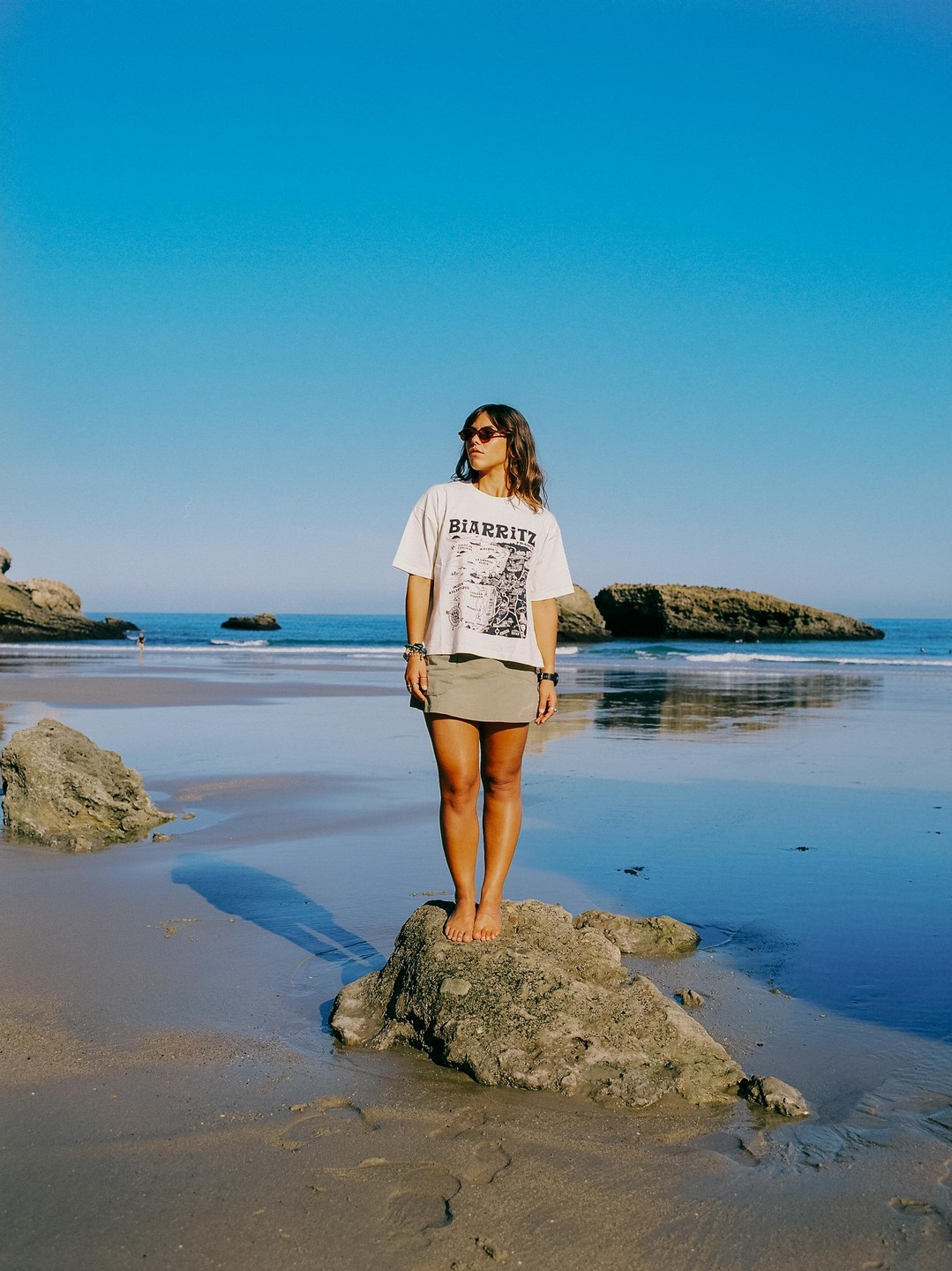 A woman stands barefoot on a rock on a beach with the ocean and other rocks in the background.