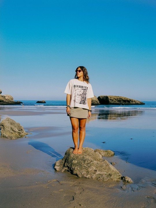 A woman stands barefoot on a rock on a beach with the ocean and other rocks in the background.