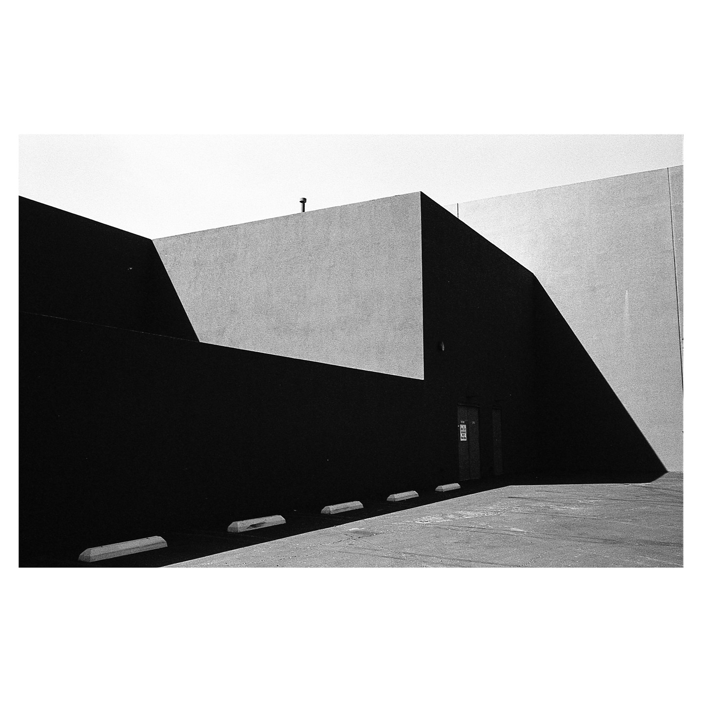 A black and white photograph captures the stark contrast of light and shadow on the angular walls of a building, with parking blocks visible in the foreground.