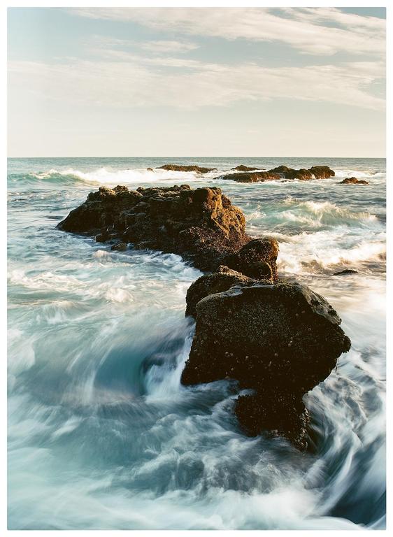 Waves crash around several large rocks in the ocean under a partly cloudy sky.