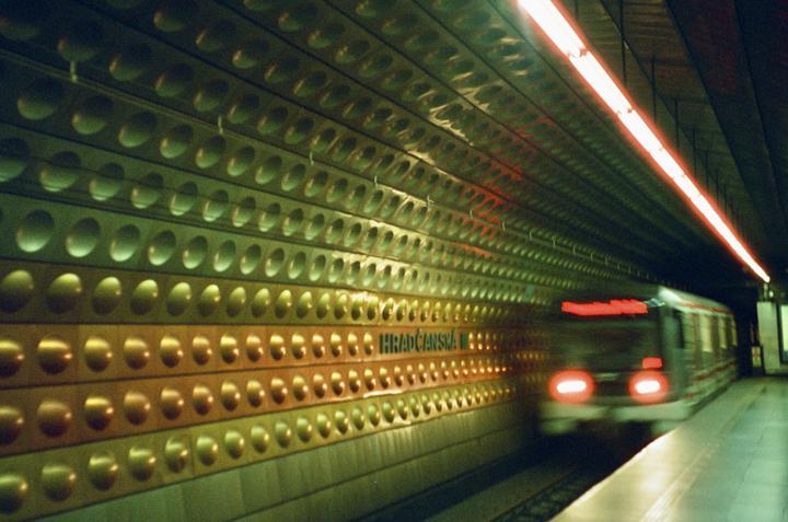 A subway train with blurred headlights approaches a station platform with a textured wall and a long red light strip on the ceiling.