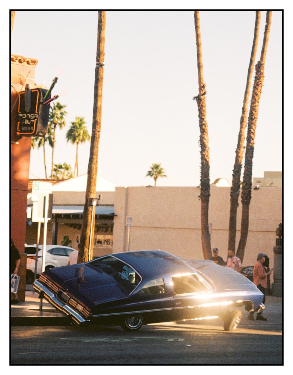 A blue lowrider car is parked on a street with its front end lifted, reflecting the golden hour sun.