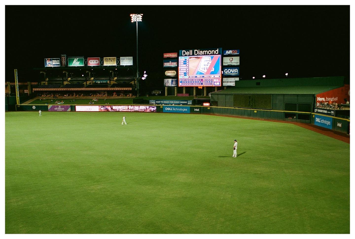A baseball field is illuminated at night with a scoreboard and advertisements visible in the background.