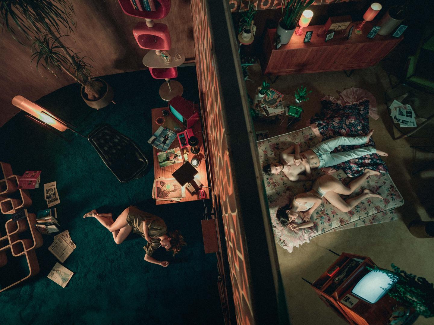 A woman lies on a blue carpet in one room while a man and a woman lie on a mattress in an adjacent room, separated by a wall.