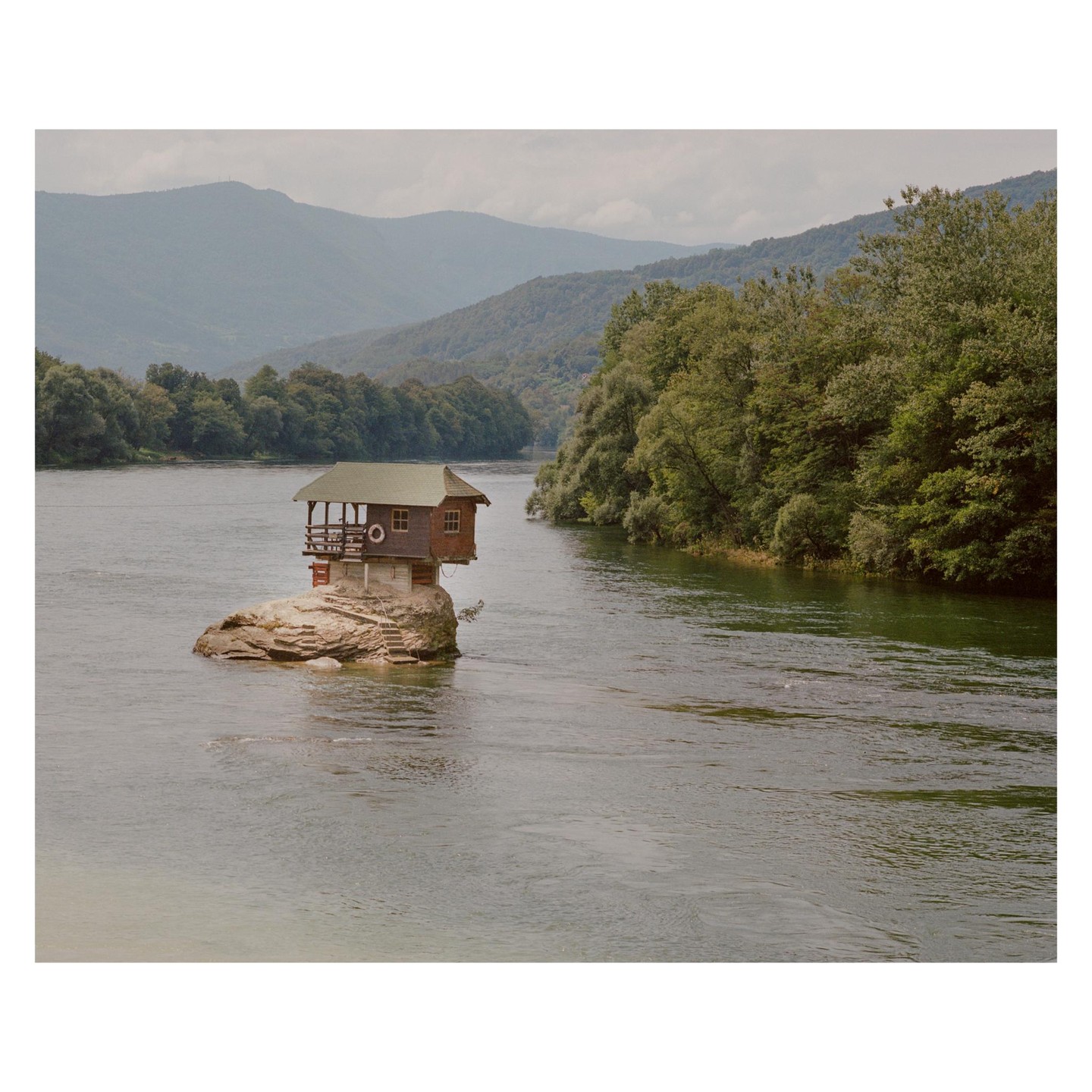 A small house sits on a rock in the middle of a river, surrounded by forested banks and distant mountains.