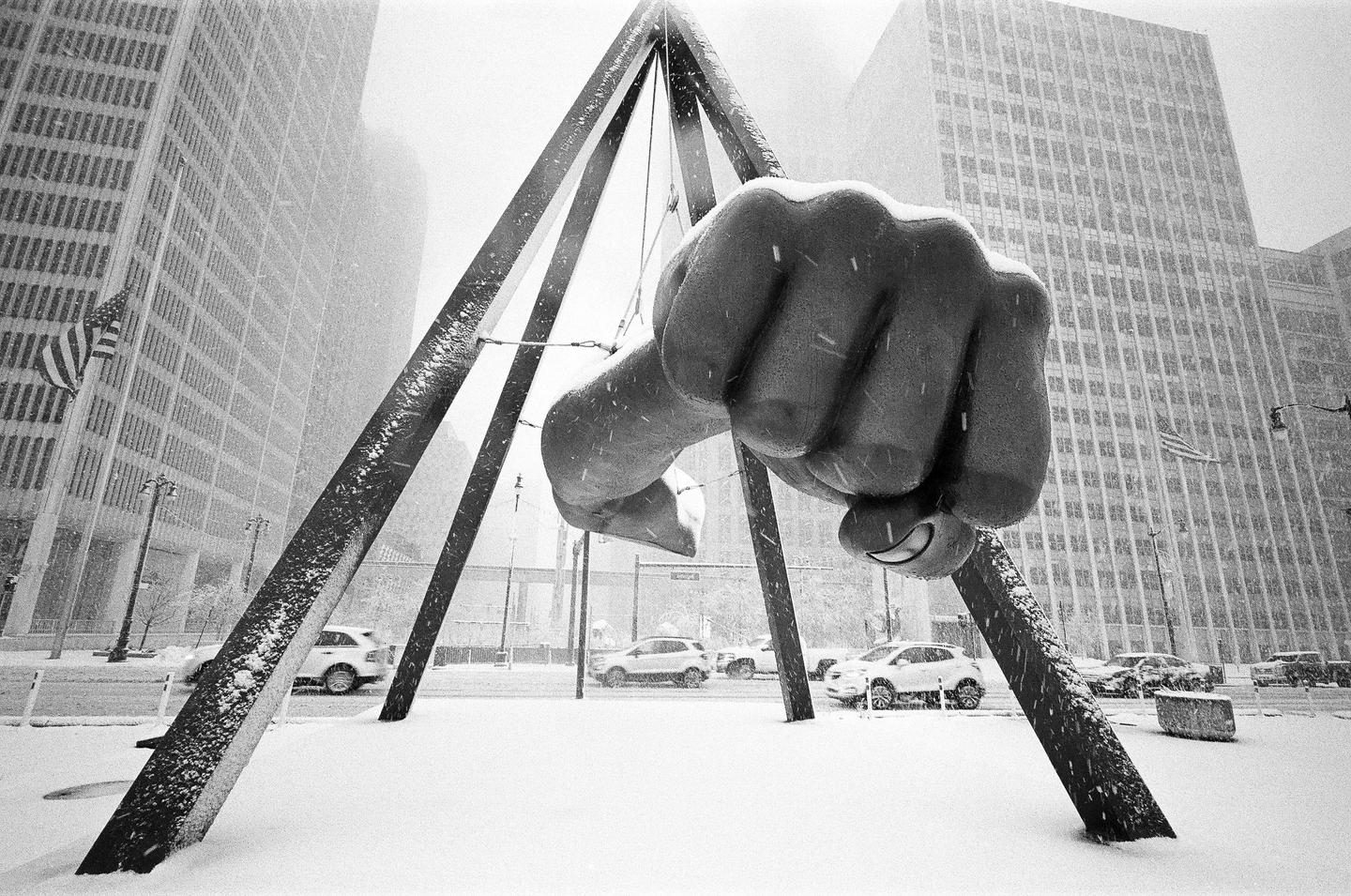 A large fist sculpture is covered in snow in a city street with tall buildings and cars in the background.