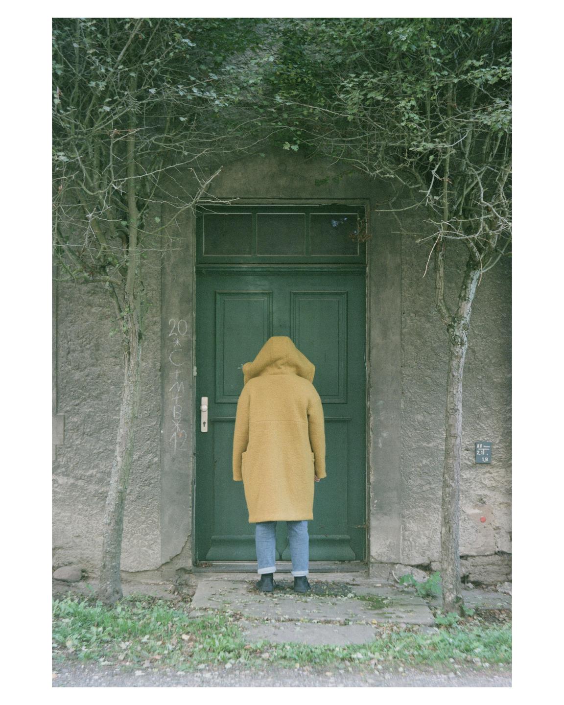 A person in a yellow coat stands facing a green door with trees on either side.